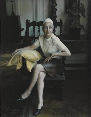 A color photograph of a seated older White woman wearing a white beanie, white sweater, gray skirt leaning toward the viewer with her elbows on the arms of the chair.