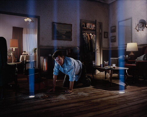 A color photograph of a White man on his hands and knees in a living room looks at beams of light coming through holes in the floor.
