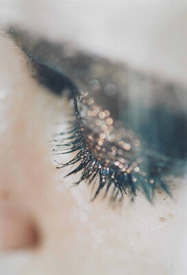 A close-up color photograph of a White woman's eyelid covered with sparkly eyeshadow and her lashes thick with mascara.