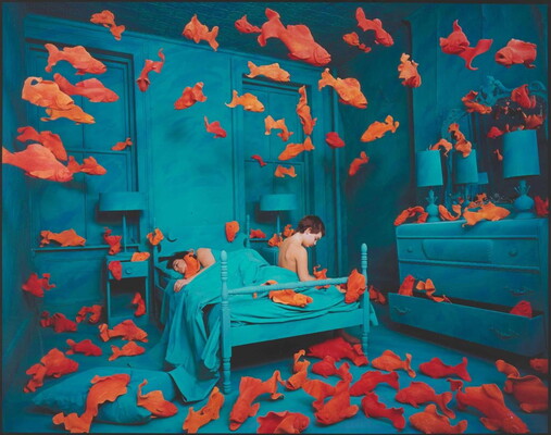 A color photograph of a bedroom with blue walls and furniture, orange goldfish floating throughout, and two White people on the bed: one sleeping and one sitting on the edge.