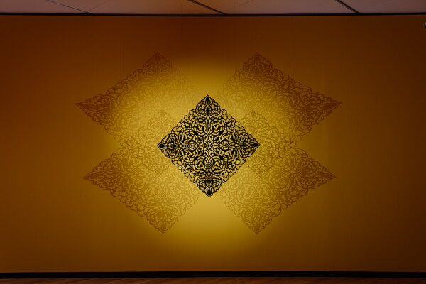 A square piece of steel with an intricate design cut into it projects ethereal shadows on a gold wall behind it.