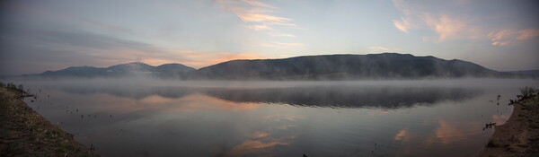 A color photograph of morning fog over a body of water with dark hills in the background.