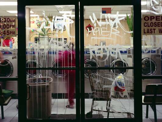 A color photograph of a laundromat at night as seen through glass doors covered in white graffiti.