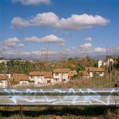 A color photograph of a graffitied guardrail with houses in the background under a cloud-filled sky.