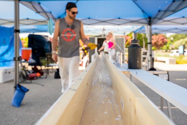 A photograph of a White man pulling a long thread of clear glass into a wood trough.
