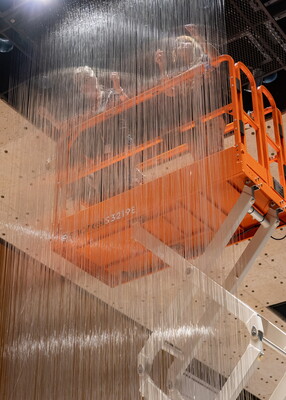 Two people stand on a lift in a Carter gallery hanging long threads of clear glass from the ceiling.