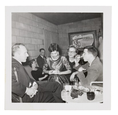 A black-and-white snapshot of formally-dressed people at a party in a cinderblock room.