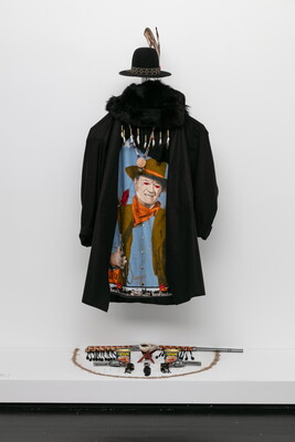 An installation view of a black open-crown cowboy hat with feather sticking up from the headband, a black coat with a fur cowl over a blue shirt with a painting of a White vampire cowboy all hung over a display of Spam, revolvers, and a rifle.