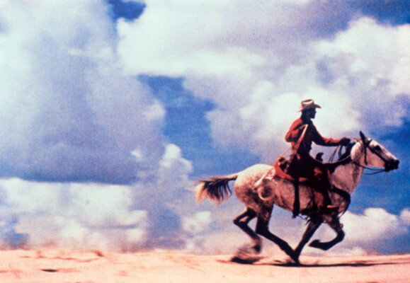 A color photograph of a man on a running horse in a dusty landscape and a sky full of white, fluffy clouds.