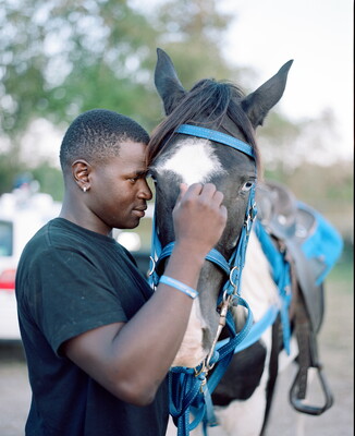 A color photograph of a Black man nuzzling and scratching the forehead of a horse wearing a blue bridle