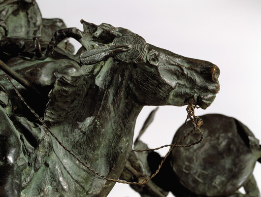 A detail of a bronze sculpture of two horses in mid-gallop, with both riders lunging toward a crouching man behind a shield.
