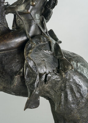 A detail of a bronze sculpture showing saddle of a man on horseback holding a piece of scalp and hair in his hand thrusted above his head triumphantly.