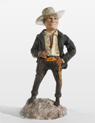 A painted sculpture of a man wearing a cowboy hat, a light-colored shirt, and dusty pants and jacket standing on a rock, smoking a cigarette.