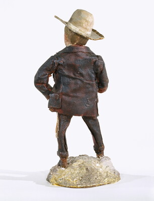 A painted sculpture of a man wearing a cowboy hat, a light-colored shirt, and dusty pants and jacket standing on a rock, smoking a cigarette.