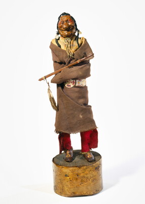 A painted wood sculpture of a smiling Native American man with exaggerated features wrapped in a blanket with a pipe resting on his arm.