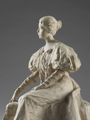 A white, plaster sculpture of a young woman sitting on a bench in a long dress with puffy sleeves.