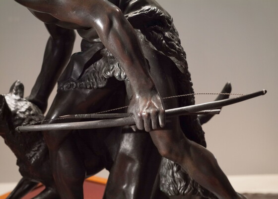 A detail of bronze sculpture of a young man holding a bow and arrow, standing hunched low with a dog beside him.
