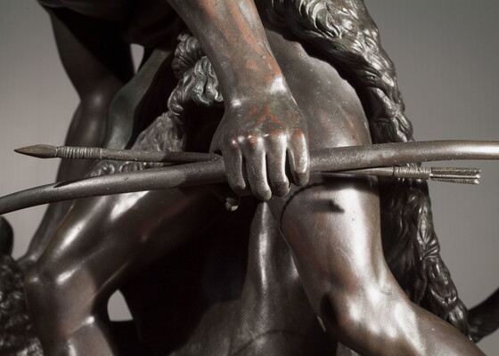 A detail of bronze sculpture of a young man holding a bow and arrow, standing hunched low with a dog beside him.