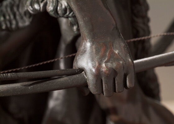 A detail of bronze sculpture of a young man holding a bow and arrow, standing hunched low with a dog beside him.