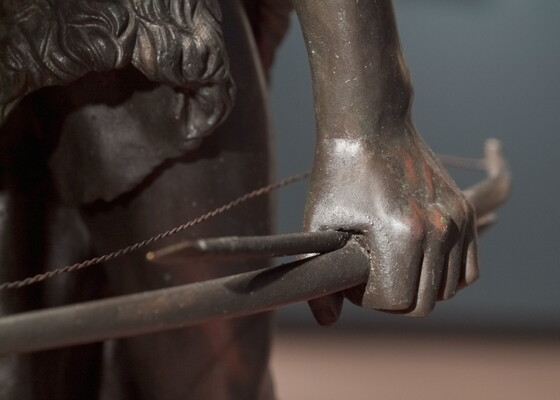 A detail of bronze sculpture of a young man holding a bow and arrow, standing hunched low with a dog beside him.