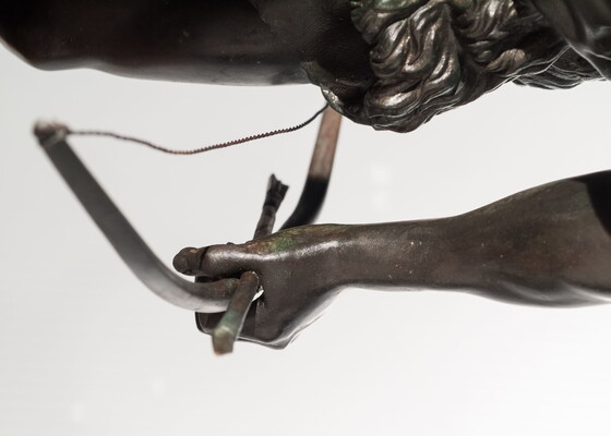 A detail of bronze sculpture of a young man holding a bow and arrow, standing hunched low with a dog beside him.