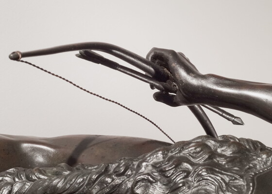 A detail of bronze sculpture of a young man holding a bow and arrow, standing hunched low with a dog beside him.