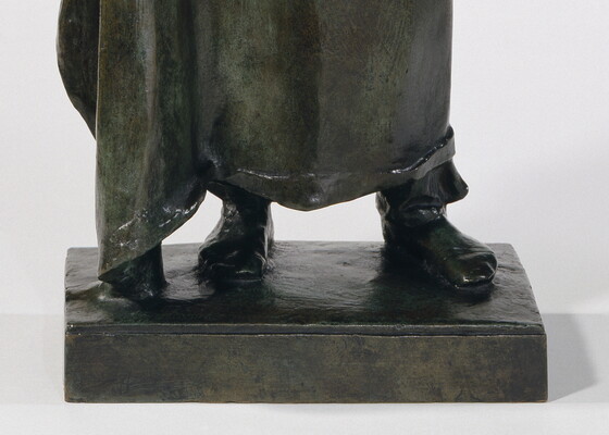 A detail of a bronze sculpture of a Native American man who stands wrapped in a blanket.