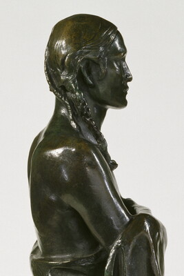 A detail of a bronze sculpture of a Native American man who stands wrapped in a blanket.