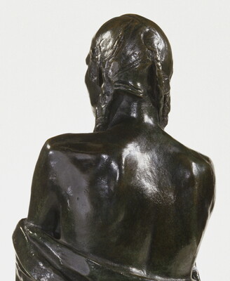 A detail of a bronze sculpture of a Native American man who stands wrapped in a blanket.