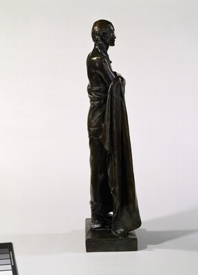 A bronze sculpture of a Native American man who stands wrapped in a blanket.