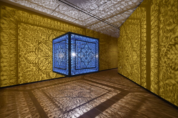 A brightly lit and suspended blue cube with laser cut shapes on all sides that cast intricate shadows on yellow walls.