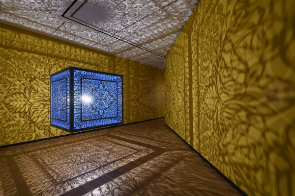 A brightly lit and suspended blue cube with laser cut shapes on all sides that cast intricate shadows on yellow walls.