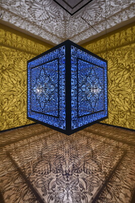 A brightly lit and suspended blue cube with laser cut shapes on all sides that cast intricate shadows on yellow walls.