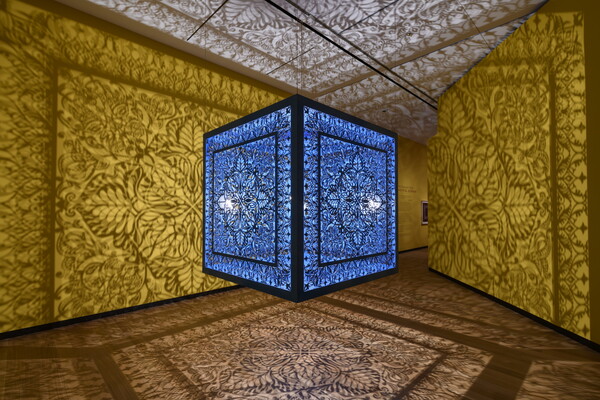 A brightly lit and suspended blue cube with laser cut shapes on all sides that cast intricate shadows on yellow walls.