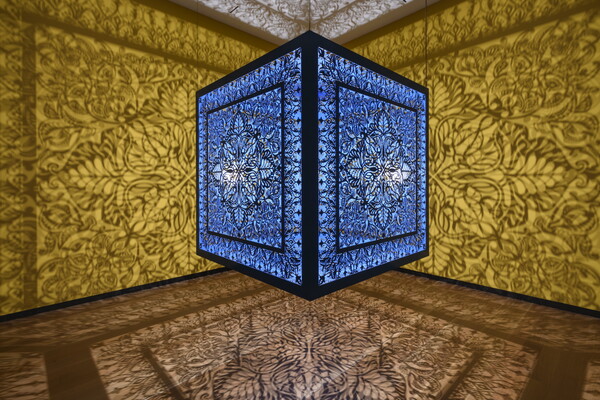 A brightly lit and suspended blue cube with laser cut shapes on all sides that cast intricate shadows on yellow walls.