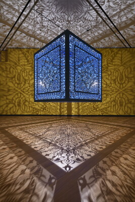 A brightly lit and suspended blue cube with laser cut shapes on all sides that cast intricate shadows on yellow walls.