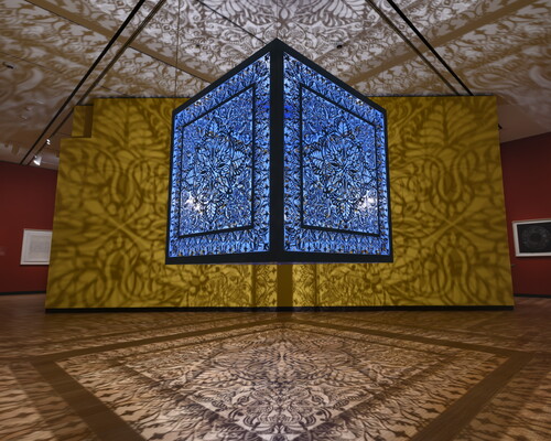 A brightly lit and suspended blue cube with laser cut shapes on all sides that cast intricate shadows on yellow walls.
