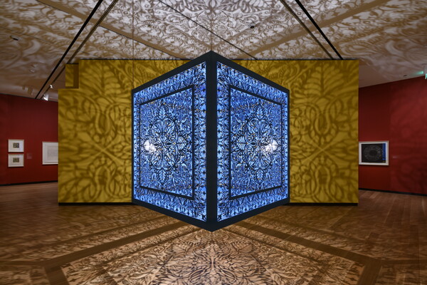 A brightly lit and suspended blue cube with laser cut shapes on all sides that cast intricate shadows on yellow walls.