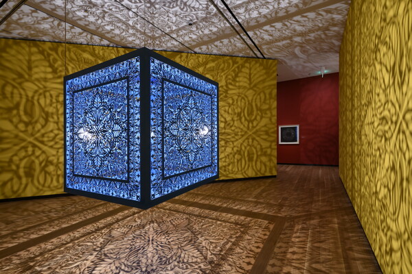 A brightly lit and suspended blue cube with laser cut shapes on all sides that cast intricate shadows on yellow walls.