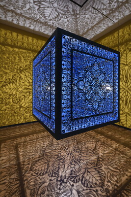 A brightly lit and suspended blue cube with laser cut shapes on all sides that cast intricate shadows on yellow walls.
