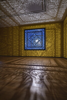 A brightly lit and suspended blue cube with laser cut shapes on all sides that cast intricate shadows on yellow walls.