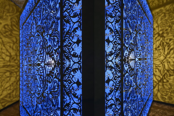 A detail of a brightly lit and suspended blue cube with laser cut shapes on all sides that cast intricate shadows on yellow walls.
