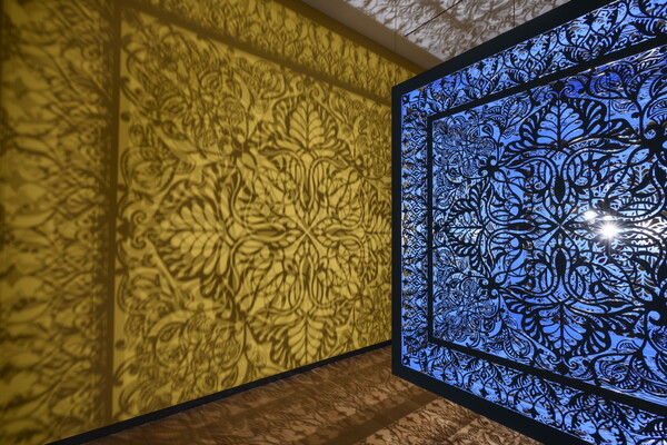 A detail of a brightly lit and suspended blue cube with laser cut shapes on all sides that cast intricate shadows on yellow walls.