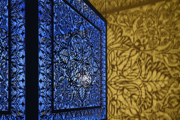 A detail of a brightly lit and suspended blue cube with laser cut shapes on all sides that cast intricate shadows on yellow walls.