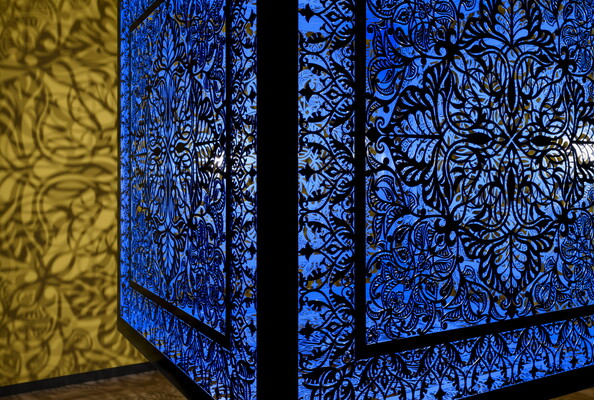 A detail of a brightly lit and suspended blue cube with laser cut shapes on all sides that cast intricate shadows on yellow walls.