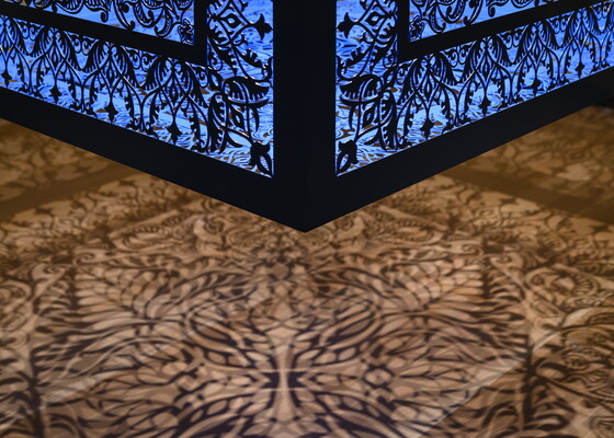 A detail of a brightly lit and suspended blue cube with laser cut shapes on all sides that cast intricate shadows on yellow walls.