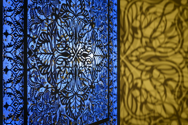 A detail of a brightly lit and suspended blue cube with laser cut shapes on all sides that cast intricate shadows on yellow walls.