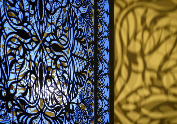 A detail of a brightly lit and suspended blue cube with laser cut shapes on all sides that cast intricate shadows on yellow walls.