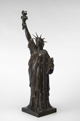 A bronze sculpture of a woman wearing a pointed crown, one arm raised holding a torch, draped in loose fabric, cradling a book in her other arm.