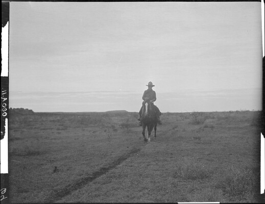A black-and-white image of a cowboy on horseback trotting on a path toward the viewer in a barren landscape.